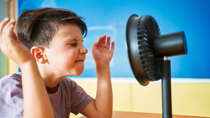 Boy refreshing his face and having fun while sitting in front of a small electric fan in the classroom at school.